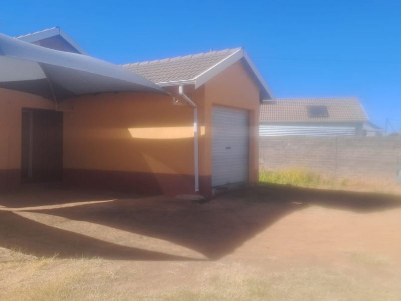 Rear view of the property featuring a roller garage door, a shade sail structure, and a surrounding boundary wall on an unpaved lot.