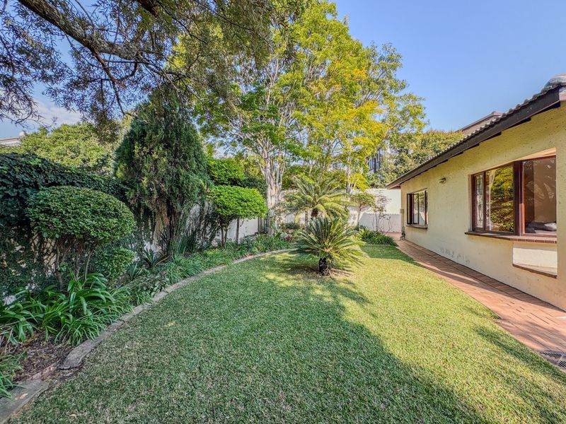 A well-maintained rear garden featuring a lush lawn, mature trees, and a brick-paved walkway adjacent to the building exterior.