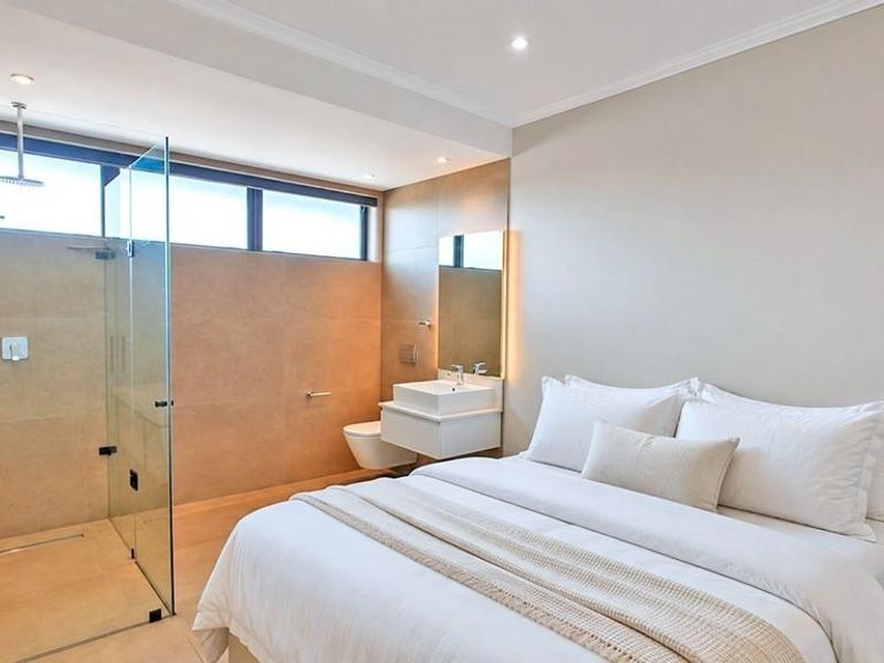 Modern bedroom featuring an open-plan en-suite bathroom with a glass-enclosed shower, wall-mounted vanity, and wall-hung toilet, finished with neutral tiling.