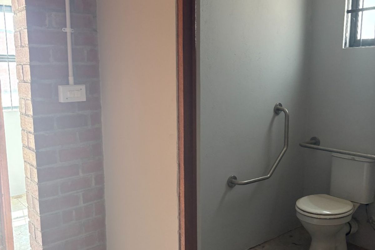 A functional bathroom space featuring a toilet, safety grab rails, and exposed brick accents, currently requiring floor finishing and cosmetic updates.