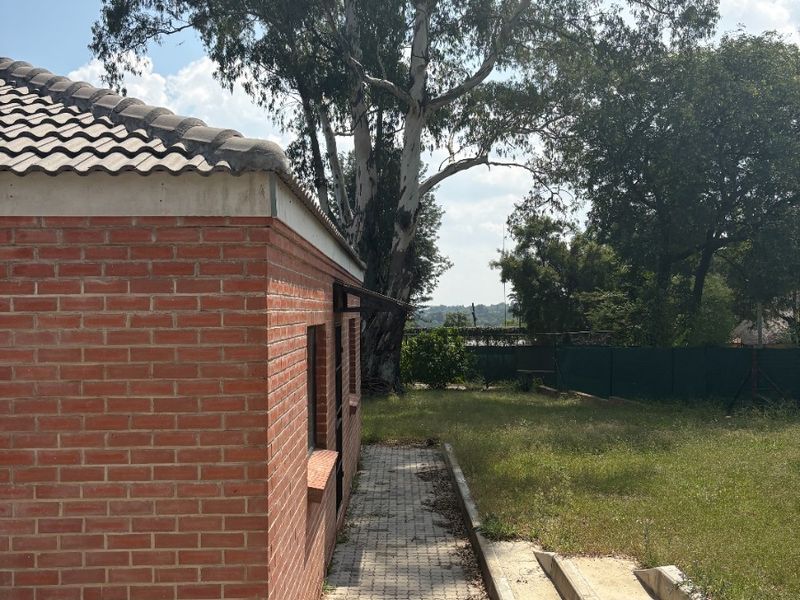 A view of the rear exterior of the brick farm building, featuring a paved walkway, concrete drainage channel, and surrounding garden area.