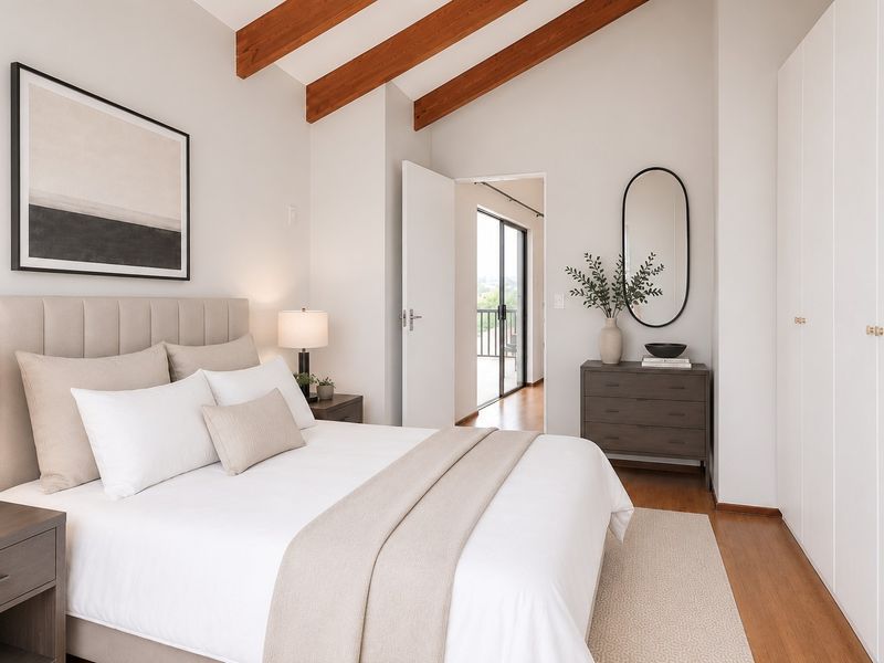 A bright, well-maintained bedroom featuring exposed timber ceiling beams, hardwood flooring, and integrated built-in wardrobes with access to an adjoining balcony.