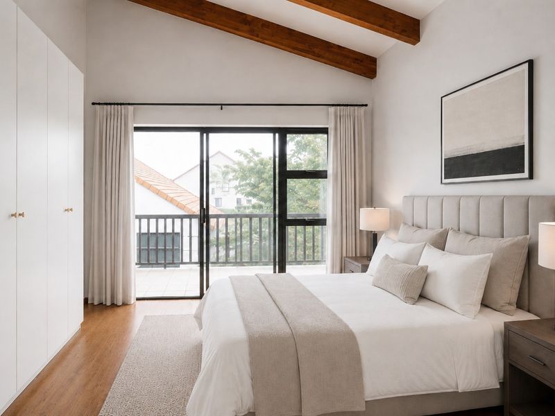A bright, modern bedroom featuring vaulted ceilings with exposed timber beams, built-in wardrobes, hardwood flooring, and direct access to a private balcony.