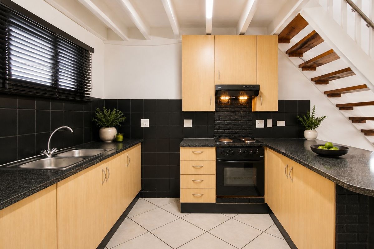 A functional kitchen featuring light wood cabinetry, black tiled backsplash, granite-look countertops, built-in oven and hob, and an adjacent staircase.