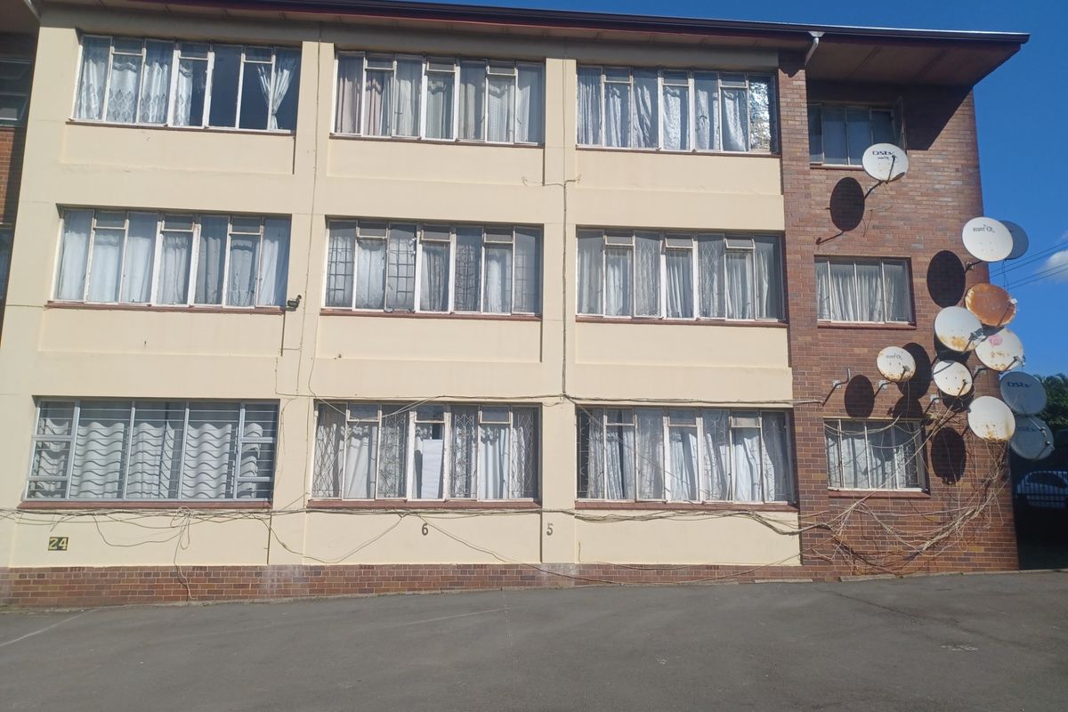 Exterior view of a multi-story apartment building featuring brick and painted facade, metal-framed windows with security bars, and multiple satellite dish installations.