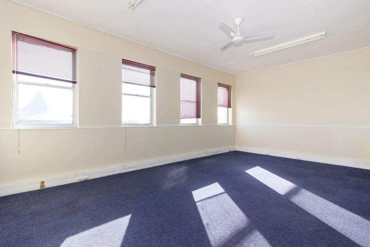 A bright, empty office space featuring blue carpet, multiple windows with venetian blinds, a ceiling fan, and neutral wall finishes.