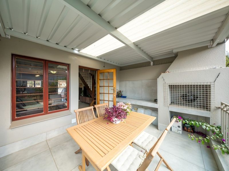 A covered patio area featuring tiled flooring, a built-in braai, and direct access to the interior through a timber-framed glass door.