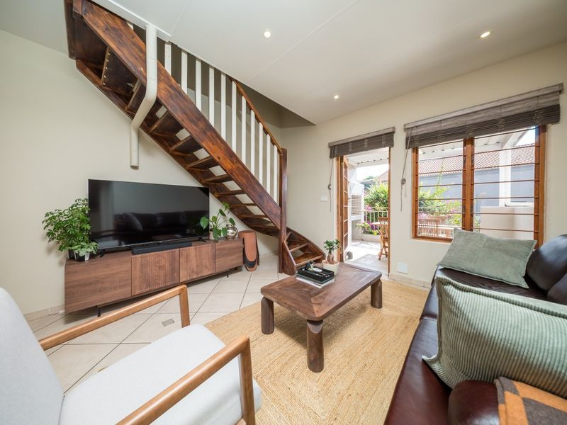 A bright living room featuring a prominent wooden staircase, tiled flooring, and direct access to an outdoor patio area through timber-framed doors.