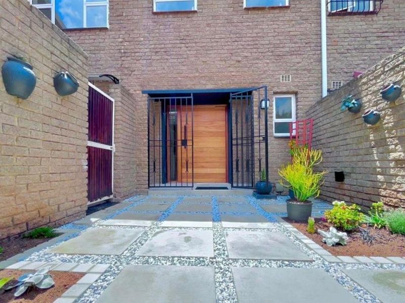 A well-maintained front entrance featuring a modern timber door, secure metal gate, and a neatly paved courtyard with brick retaining walls.