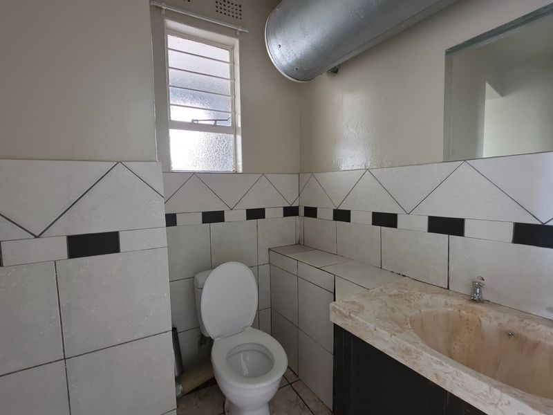 A functional bathroom featuring a toilet, vanity with basin, and tiled walls. The space includes an exposed ventilation duct and a louver window.