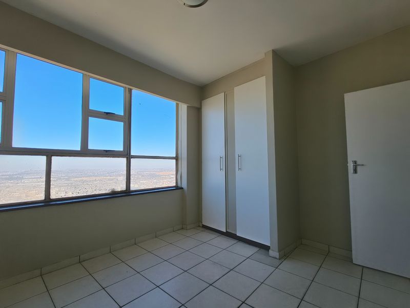 A bright, empty bedroom featuring tiled flooring, built-in cupboards, and a large window offering expansive elevated views.