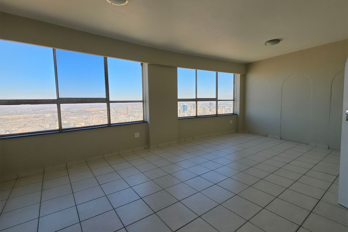 An empty, sunlit living area featuring tiled flooring and expansive windows offering panoramic city views.