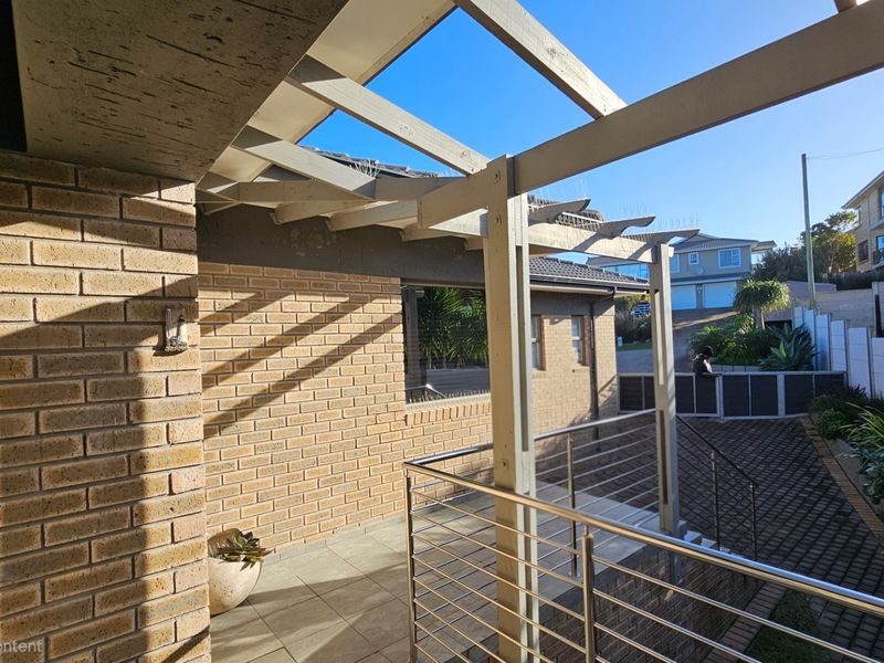 An elevated outdoor patio area featuring a timber pergola, stainless steel balustrade, and tiled flooring, overlooking the paved driveway and front garden.