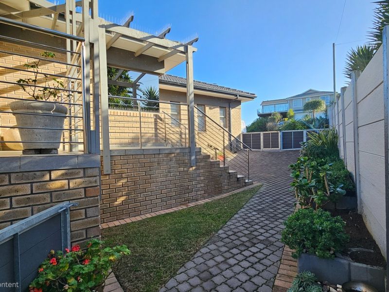 A well-maintained rear exterior area featuring a brick facade, paved driveway, and a pergola structure with stairs leading to the main dwelling.