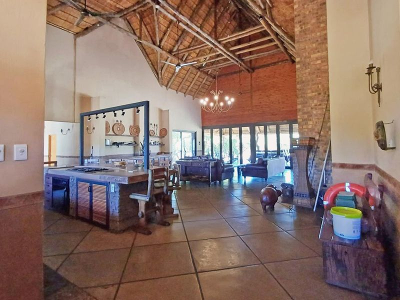 Spacious open-plan living area featuring a high exposed thatch ceiling, brick fireplace, and a central kitchen island with tiled flooring throughout.