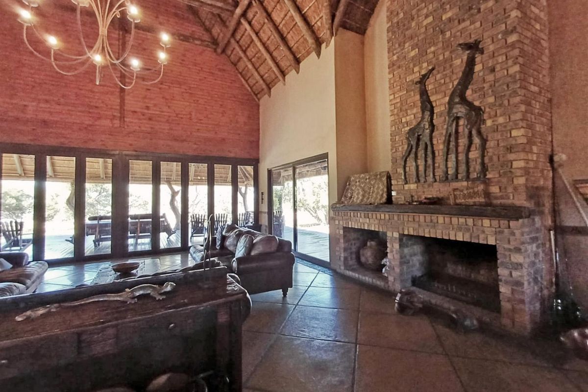 Spacious living room featuring a high exposed thatch ceiling, large brick fireplace, tiled floors, and expansive glass doors opening to an outdoor patio area.