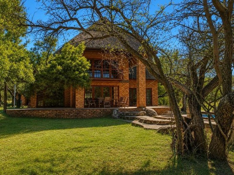 Rear view of a double-story brick house featuring a thatched roof, a spacious covered patio, and a balcony overlooking the garden.