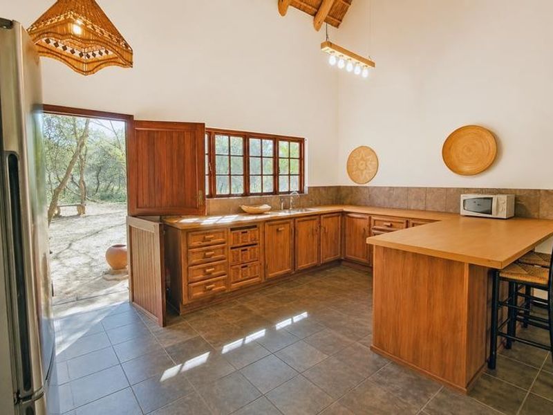 A rustic kitchen featuring wooden cabinetry, tiled flooring, a breakfast counter, and a vaulted ceiling with exposed timber beams, opening directly to the outdoors.