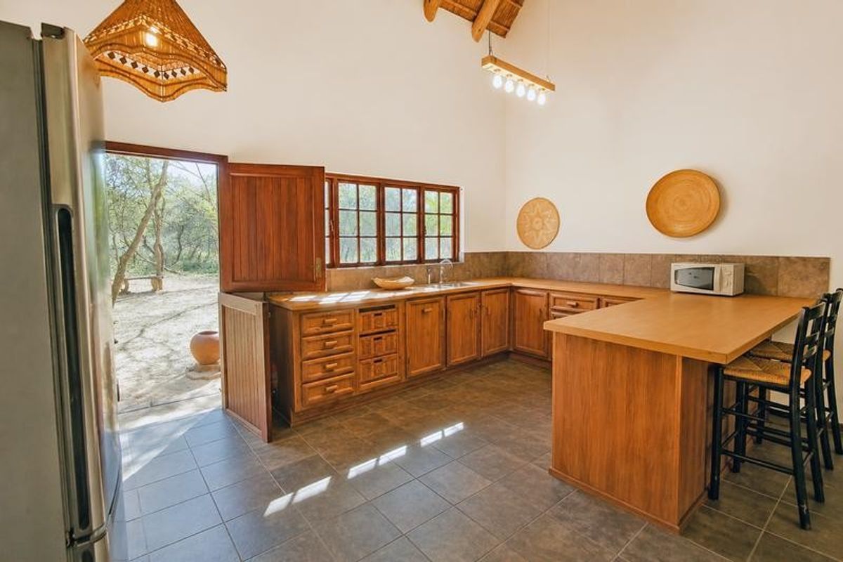A rustic kitchen featuring wooden cabinetry, tiled flooring, a breakfast counter, and a vaulted ceiling with exposed timber beams, opening directly to the outdoors.