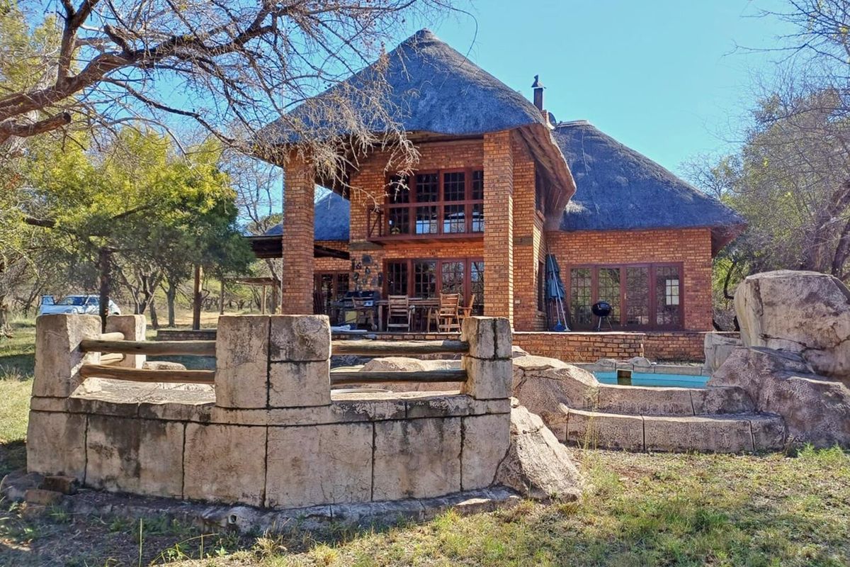 Rear view of a double-story brick house with a traditional thatch roof, featuring a swimming pool, stone retaining wall, and a timber balcony.