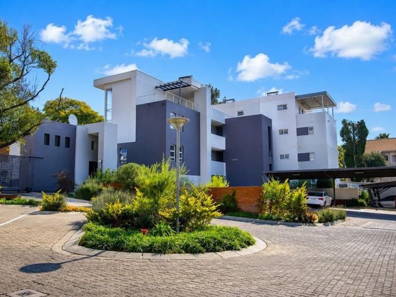 A view of the modern apartment complex exterior, featuring a paved driveway, landscaped garden area, and covered parking facilities.