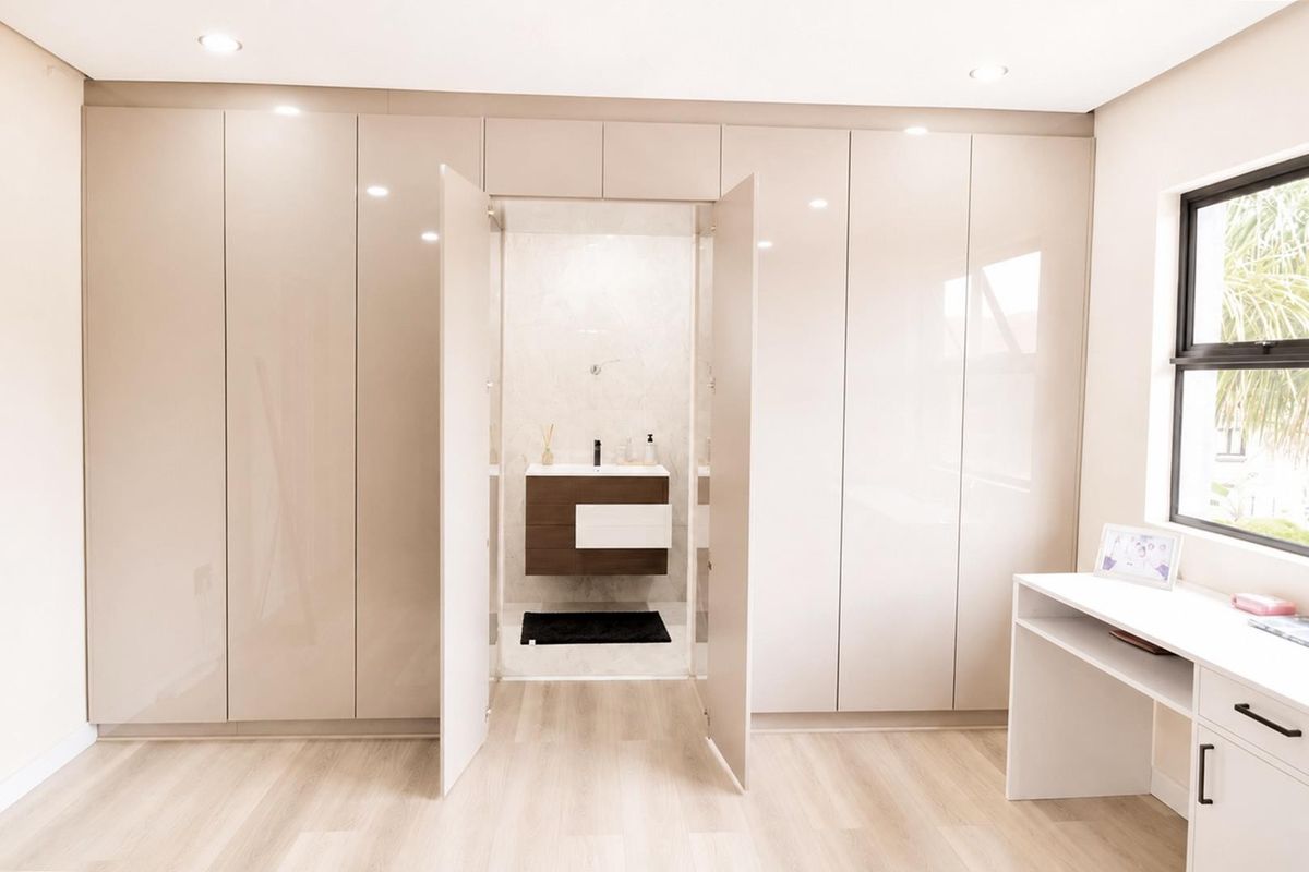 Modern bedroom featuring extensive high-gloss built-in cupboards, integrated vanity area, and a seamless transition to an en-suite bathroom with a floating vanity.