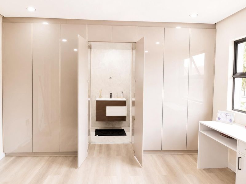 Modern bedroom featuring extensive high-gloss built-in cupboards, integrated vanity area, and a seamless transition to an en-suite bathroom with a floating vanity.