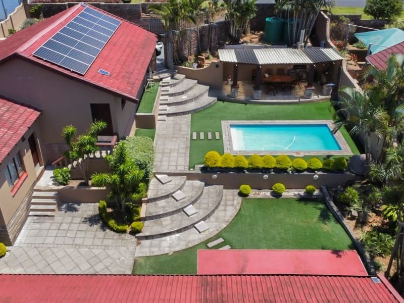 Aerial view of a residential property featuring a swimming pool, covered patio, landscaped garden, paved terrace areas, and roof-mounted solar panels.