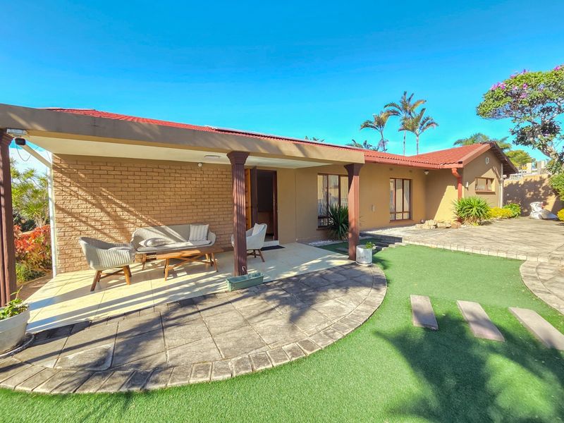 Rear view of the house featuring a covered patio area, paved flooring, and a well-maintained artificial lawn in the garden.