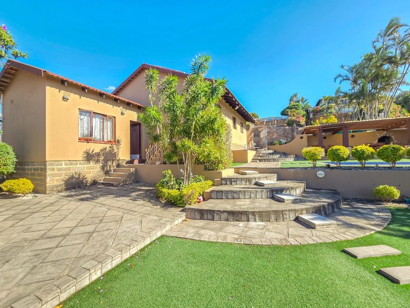 A well-maintained rear exterior featuring a paved patio, tiered stone landscaping, artificial lawn, and a covered pergola area.