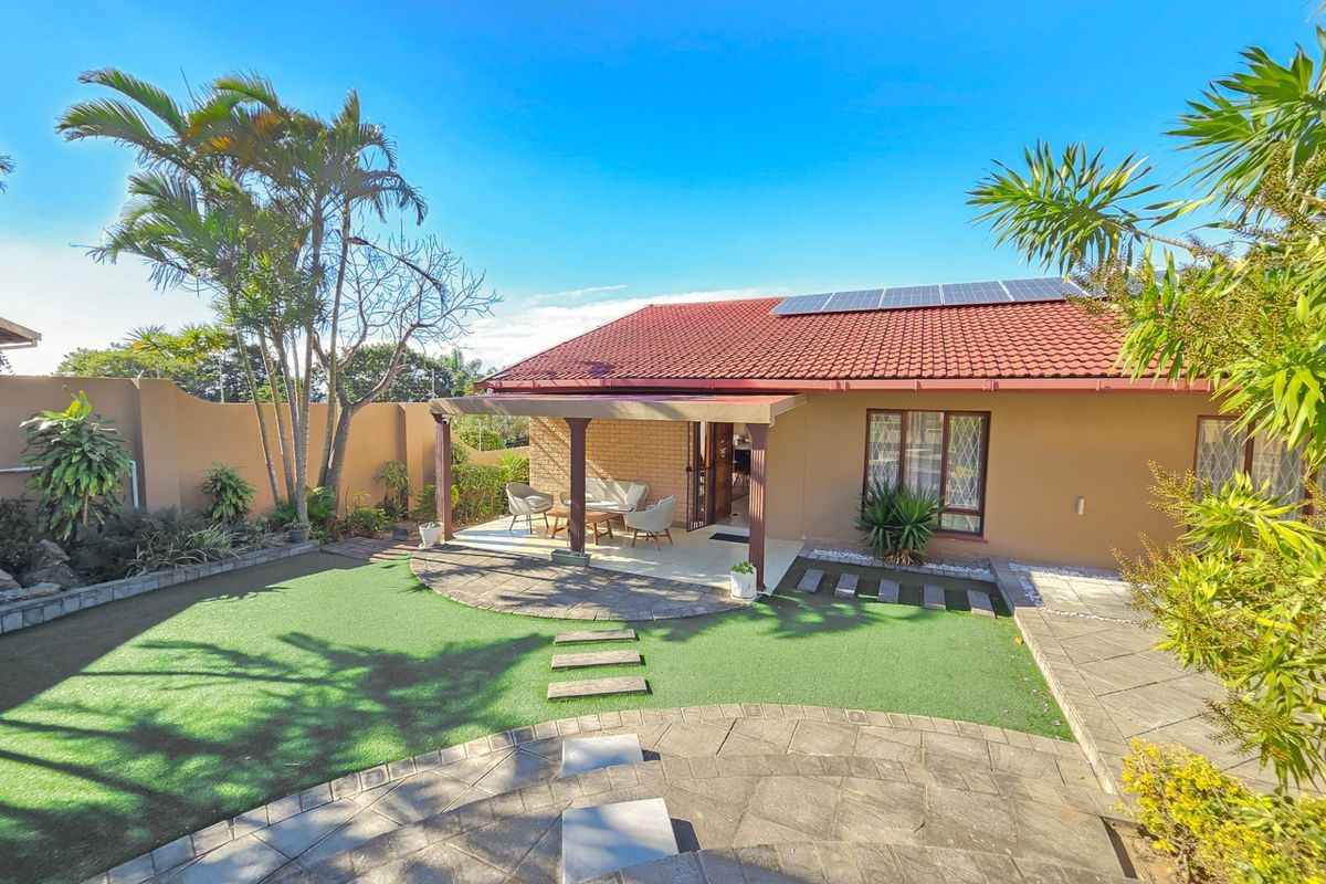 Rear view of the property featuring a covered patio, artificial lawn, paved garden pathways, and roof-mounted solar panels.