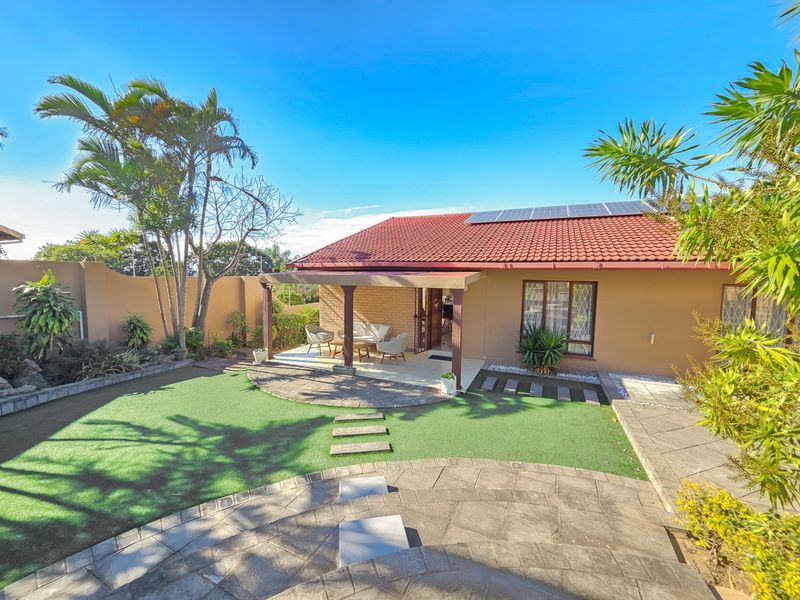 Rear view of the property featuring a covered patio, artificial lawn, paved garden pathways, and roof-mounted solar panels.