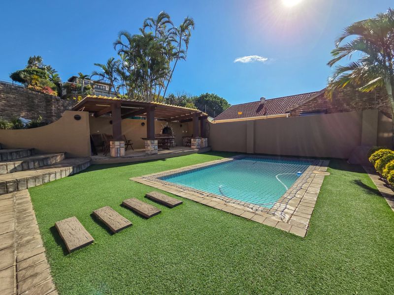 Outdoor entertainment area featuring a swimming pool with safety net, artificial lawn, and a covered patio with a built-in braai.