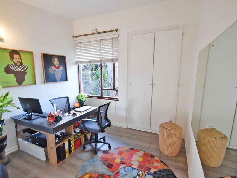 A functional home office featuring laminate flooring, a built-in wardrobe, and a window with venetian blinds, providing a bright and practical workspace.