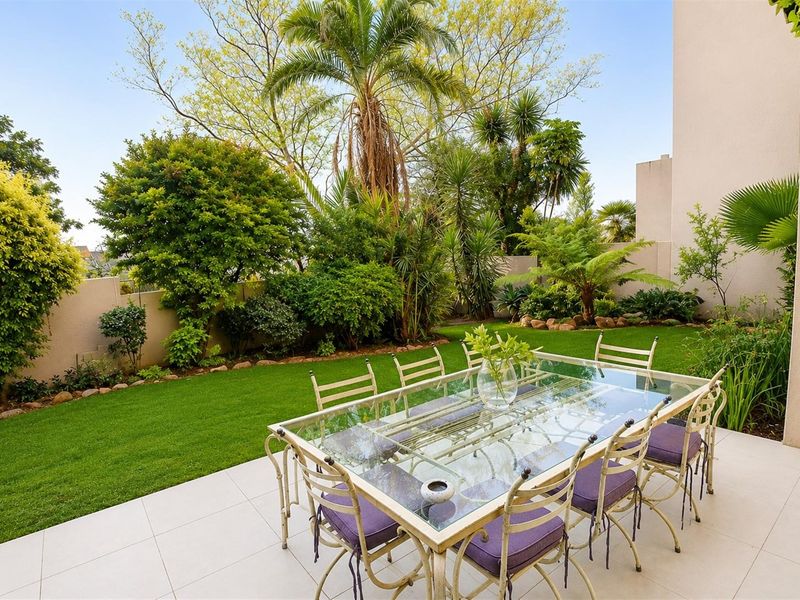 A well-maintained tiled patio area overlooking a lush, landscaped private garden with mature trees and a boundary wall.