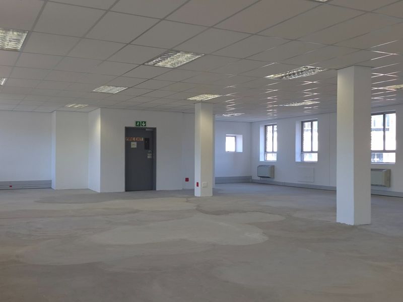 A spacious, open-plan commercial office area featuring a suspended ceiling, recessed lighting, structural columns, and wall-mounted HVAC units with unfinished concrete flooring.