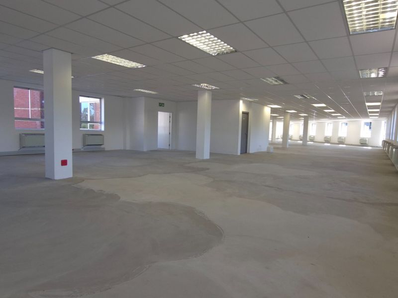 A large, open-plan commercial office space featuring a suspended ceiling, recessed lighting, structural columns, and wall-mounted HVAC units with unfinished concrete flooring.