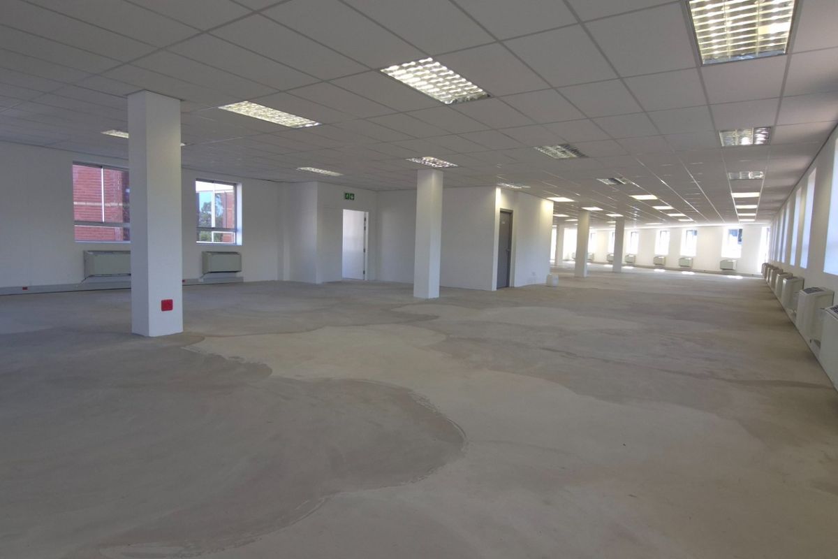 A large, open-plan commercial office space featuring a suspended ceiling, recessed lighting, structural columns, and wall-mounted HVAC units with unfinished concrete flooring.