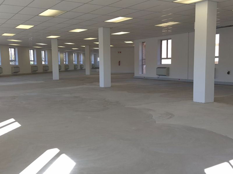 A large, open-plan retail or office space featuring a suspended ceiling, recessed lighting, structural columns, and wall-mounted HVAC units with unfinished concrete flooring.