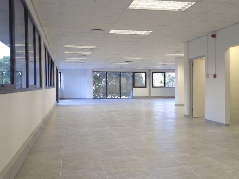 A spacious, open-plan commercial office area featuring modern tiled flooring, suspended ceilings with recessed lighting, and large windows providing ample natural light.