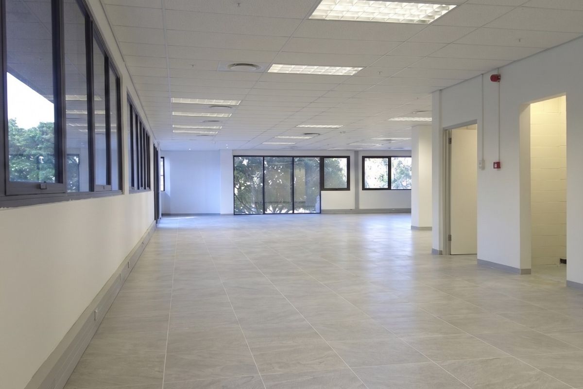 A spacious, open-plan commercial office area featuring modern tiled flooring, suspended ceilings with recessed lighting, and large windows providing ample natural light.