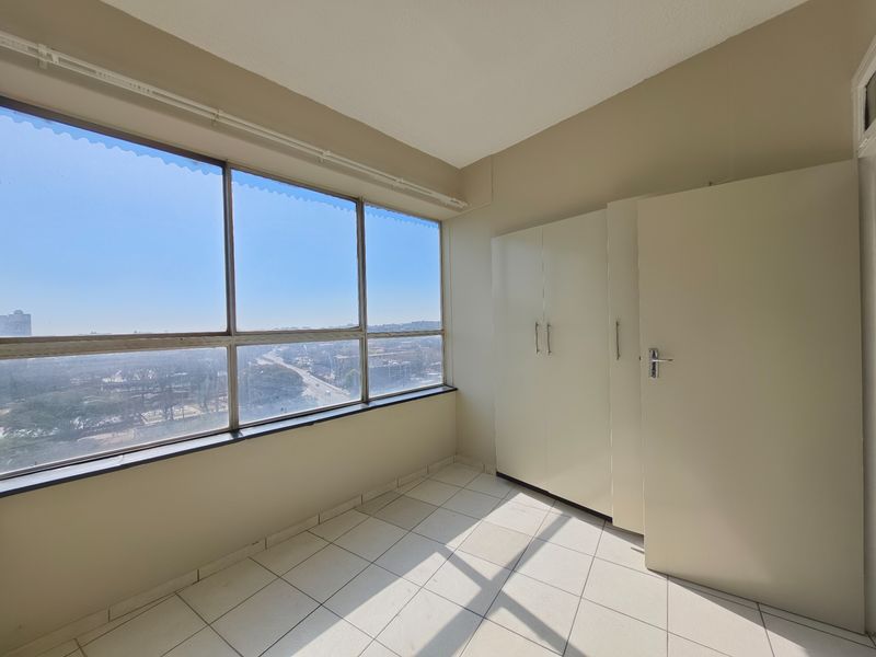 A bright, empty bedroom featuring tiled flooring, built-in cupboards, and large windows offering an expansive city view.