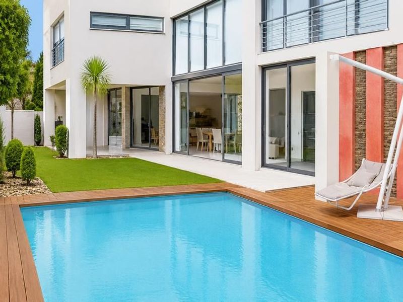 Modern rear exterior featuring a swimming pool with timber decking, artificial lawn, and large sliding glass doors leading into the home.