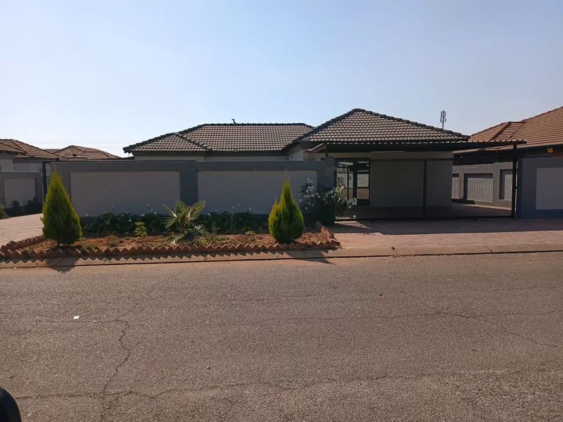 Front view of a modern single-story house featuring a tiled roof, paved driveway, and a secure perimeter wall with a gated entrance.