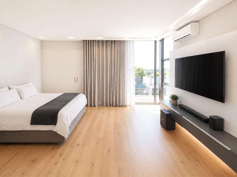 Modern bedroom featuring light hardwood flooring, recessed lighting, a wall-mounted air conditioner, and floor-to-ceiling windows with access to a balcony.