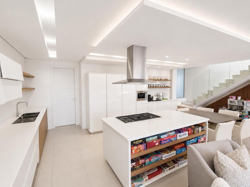 Modern open-plan kitchen featuring a central island with gas cooktop, sleek white cabinetry, integrated appliances, and a contemporary staircase with glass balustrade.