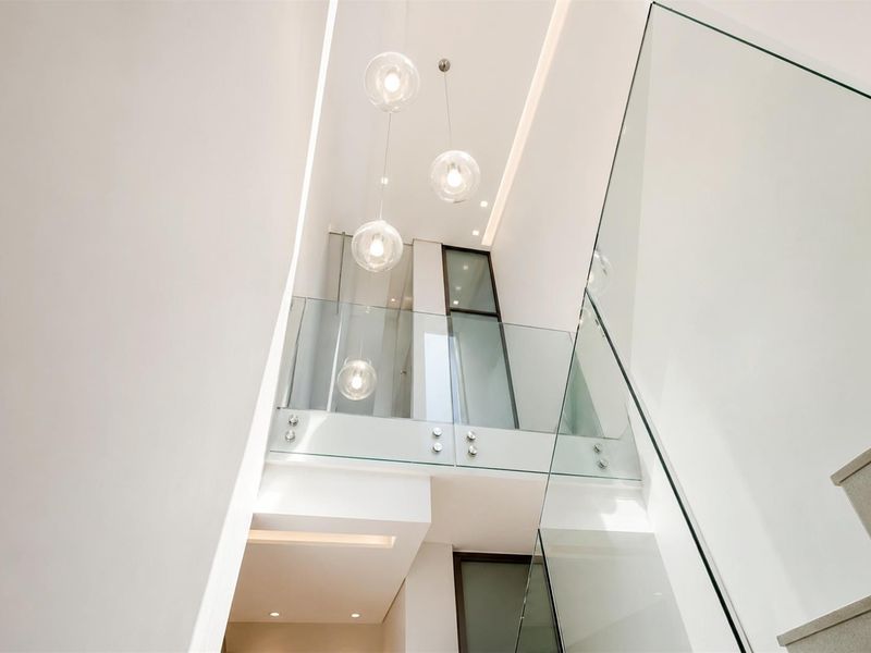 A modern, high-ceilinged hallway featuring a glass balustrade, minimalist design, and elegant pendant lighting, creating a bright and spacious architectural transition area.