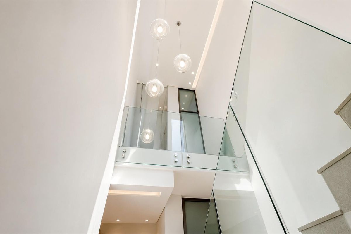 A modern, high-ceilinged hallway featuring a glass balustrade, minimalist design, and elegant pendant lighting, creating a bright and spacious architectural transition area.