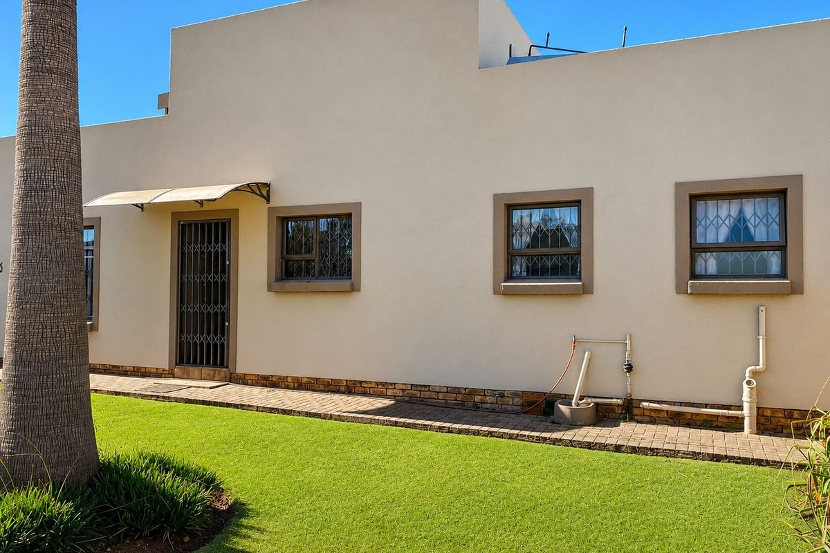Rear exterior view of the townhouse featuring a paved walkway, manicured lawn, security gates, and window bars.