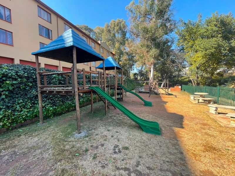Communal garden area featuring a wooden playground structure, stone seating, and a brick braai facility set against the apartment building exterior.